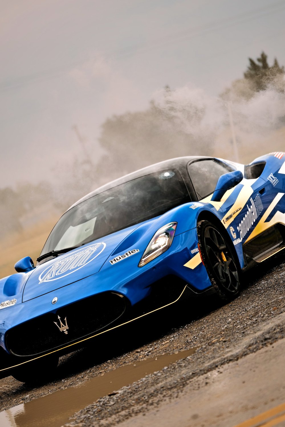 A blue Cannonball Maserati MC20 kicks up tyre smoke on a gravel shoulder
