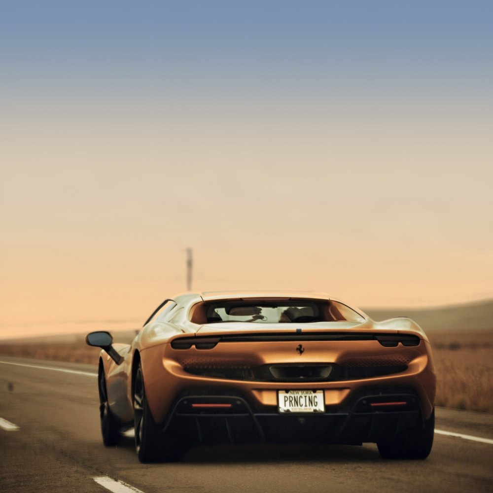 A bronze Ferrari pulls away down a two-lane desert highway at golden hour