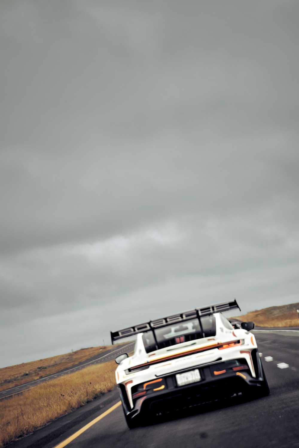 A white Porsche GT3 RS runs a two-lane highway under a wide grey sky