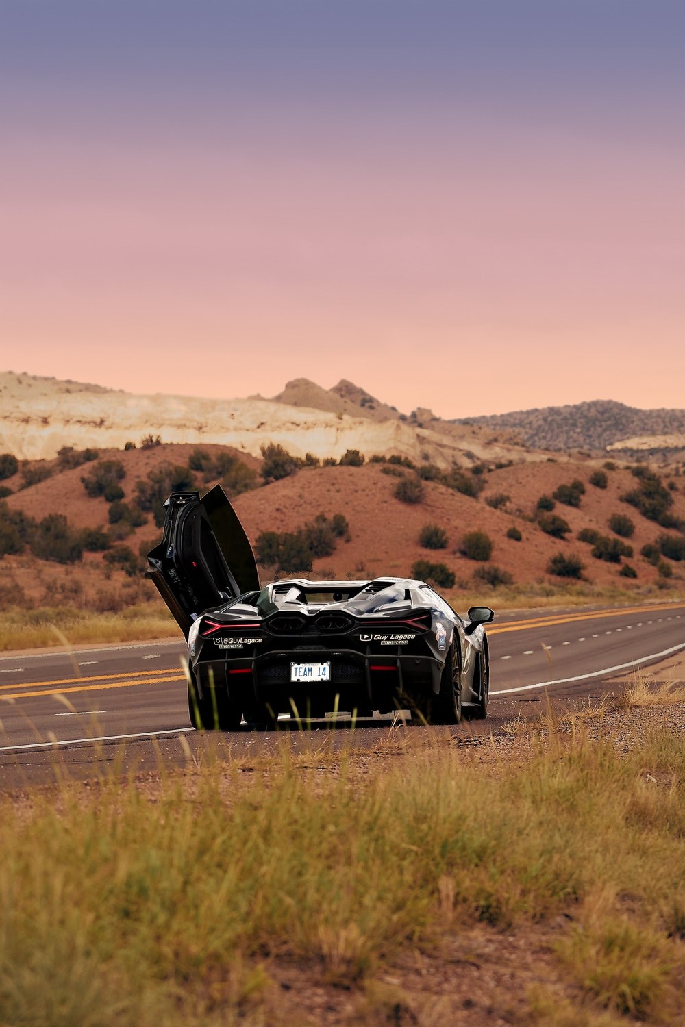 A supercar with its doors raised waits on a desert road under a pink afterglow