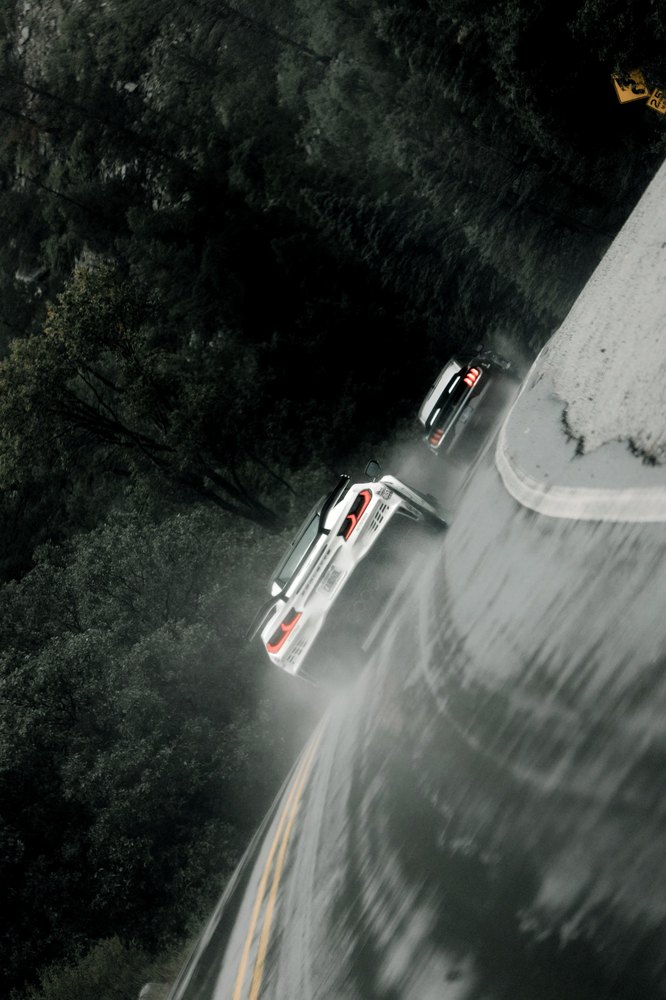 From above, two cars climb a rain-soaked mountain road through the trees
