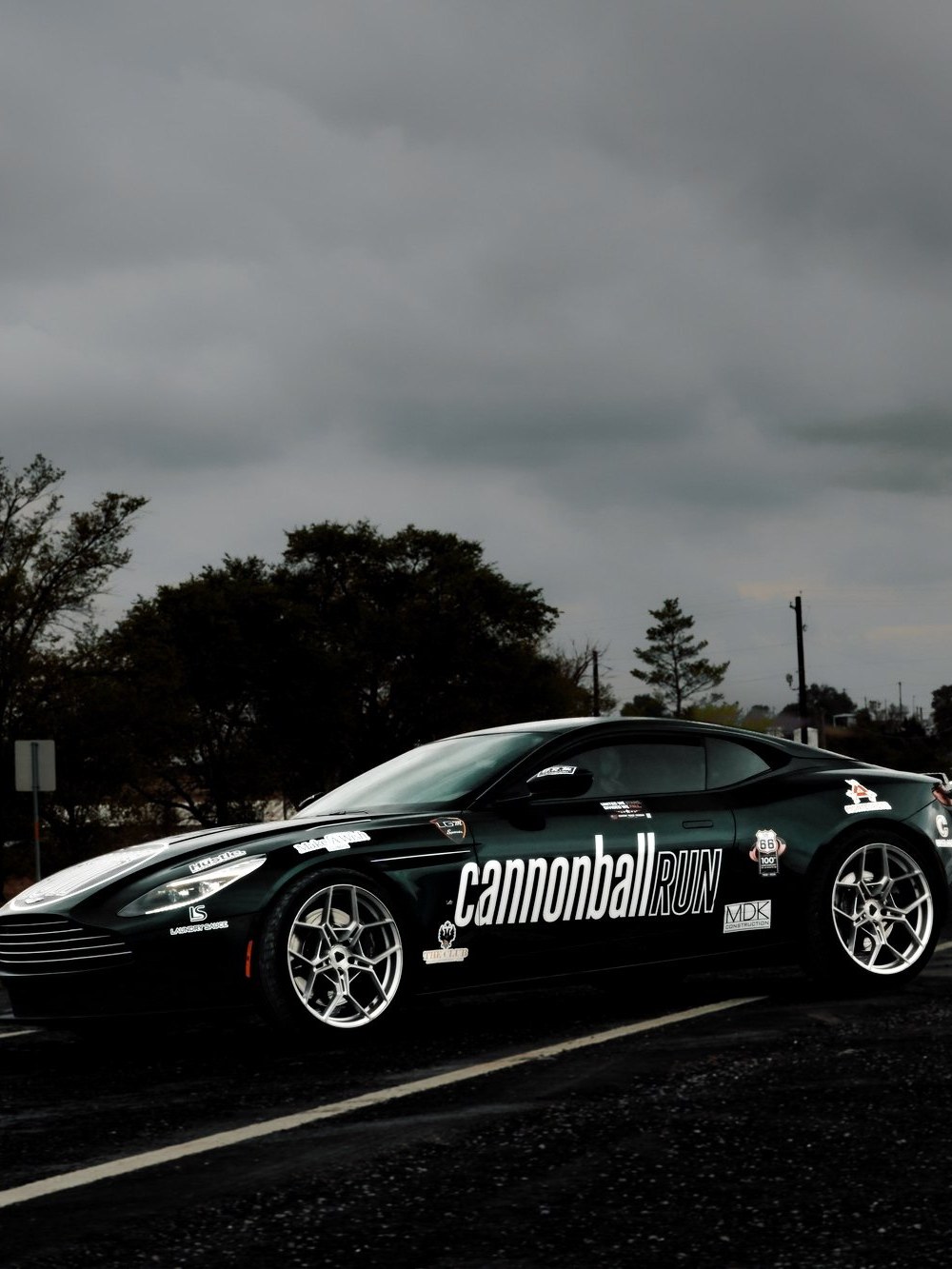 A dark grand tourer in Cannonball livery on a wet road under a heavy grey sky