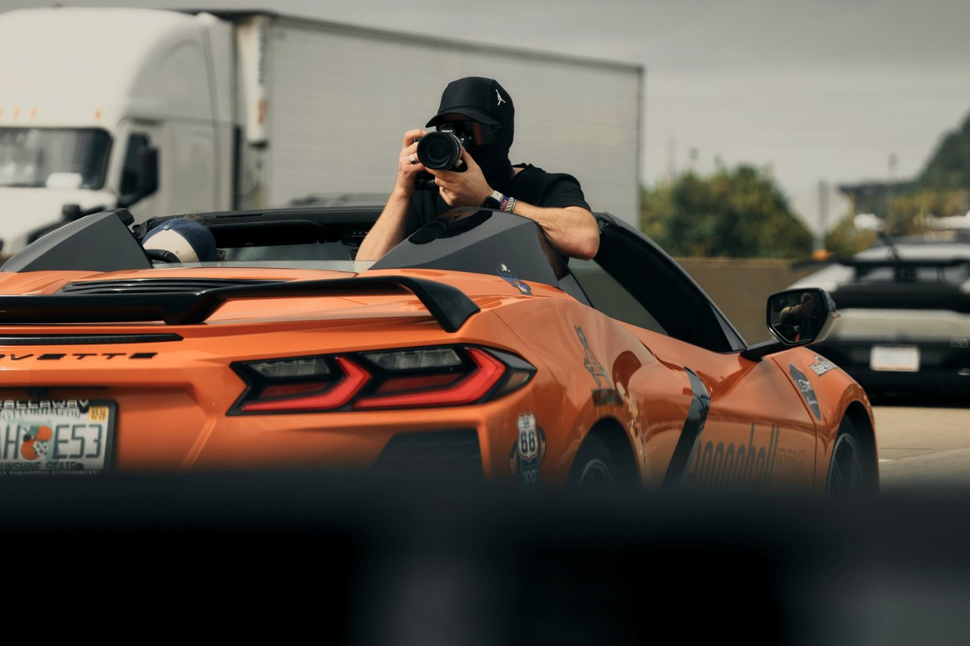 A photographer in a balaclava leans out of an orange Corvette, camera up, in traffic