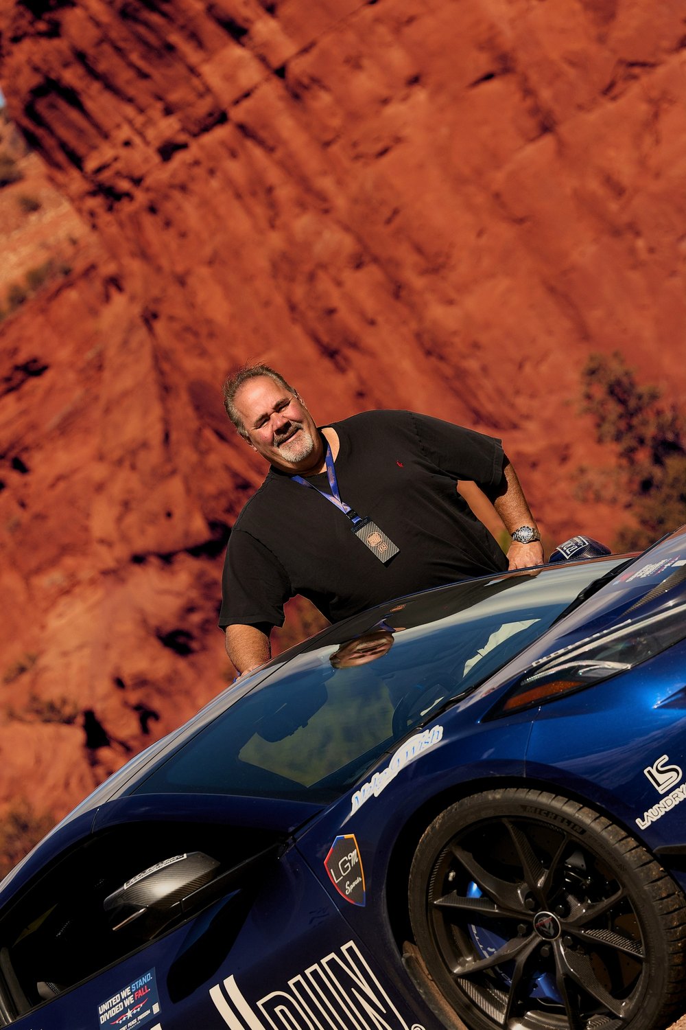 A driver leans on his blue Cannonball car beneath red rock