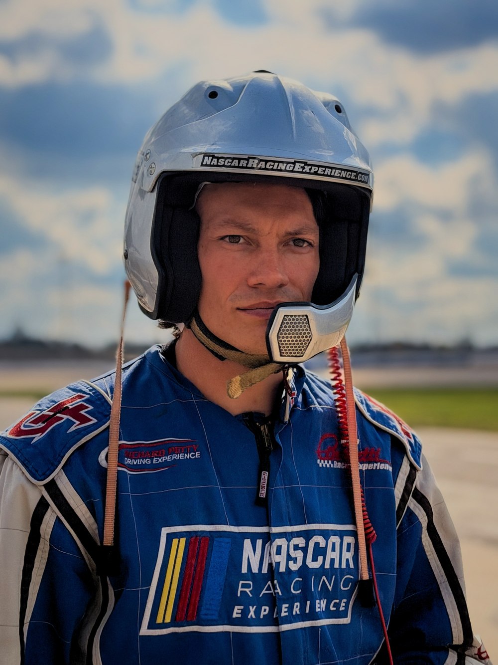 A driver in a silver helmet and blue race suit under a cloudy sky